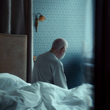 Un hombre sentado en la cama en una habitación mirando hacia otro lado, absorto en sus pensamientos.